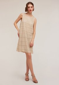 Beige ärmelloses Kleid mit einem strukturierten Wellendesign, verziert mit Perlenakzenten. Der fließende Stoff fällt sanft und endet über dem Knie.