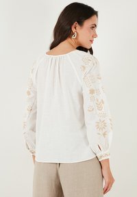 Blusa di lino bianco con ricami floreali beige sulle maniche, scollo rotondo e maniche a palloncino con dettaglio di polsino elastico.