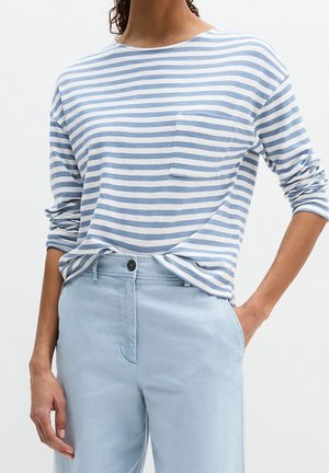 Femme portant une chemise à manches longues rayée bleu clair avec poche, rentrée dans un pantalon bleu clair taille haute, main dans la poche.