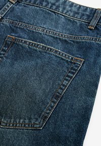 Jeans din denim, de culoare albastru închis, cu un stitching vizibil de culoare portocalie și un singur buzunar pe spate, cu o formă curbată. Textură fină.