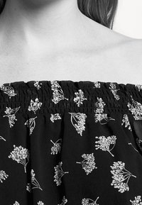 Schwarzes Off-Shoulder-Oberteil mit gerafftem elastischem Ausschnitt, das mit einem weißen Blumendruck aus kleinen Blumen und schlanken Stielen versehen ist.