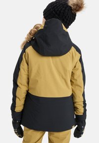 Geel en zwarte skijas met een capuchon, met een gladde textuur en contrasterende kleurvakken. Hand in gemusterde handschoenen.