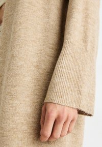 Beige stickad cardigan med en mjuk textur, ribbade ärmar och en avslappnad passform som framhäver de subtila variationerna i garnets färg.
