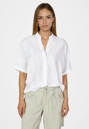 Frau mit glattem braunem Haar trägt ein weißes kurzärmeliges Hemd mit Knöpfen und hellbeige Shorts, steht vor einem schlichten Hintergrund.