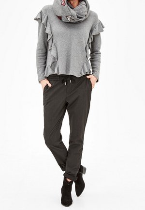 Personne portant un haut gris à manches longues avec volants et écharpe brodée, pantalon noir à cordon de serrage, et bottines noires, debout les mains dans les poches.