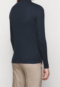 Top a maniche lunghe a coste blu navy con colletto alto, caratterizzato da un design aderente e una texture liscia. Indossato con pantaloni beige.