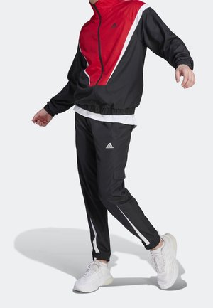 Persona vistiendo un chándal Adidas negro y rojo con rayas blancas y zapatillas blancas, posando contra un fondo liso y claro.