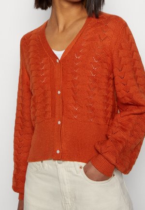 Femme portant un cardigan en maille orange boutonné avec un motif coquille sur une chemise blanche, associé à un pantalon taille haute couleur ivoire.