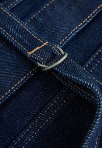 Nærbillede af mørkeblå denimstof med beige syninger og en metalring, der er løbet gennem en denimrem.
