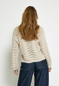 Knitted cream sweater with a wavy pattern, crew neckline, and ribbed cuffs. Paired with high-waisted dark denim pants.