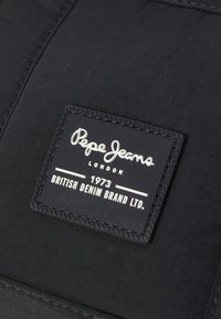 Juodos medžiagos pleistras su baltais iškiliais užrašais "Pepe Jeans London 1973 British Denim Brand Ltd." ir siūlų apvadu.
