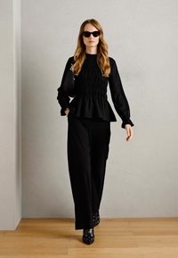 Černý smockovaný top s dlouhým rukávem a peplum pasem, spárovaný s volnými černými kalhotami a černými kotníkovými botami s hroty. Modelka má na sobě sluneční brýle.