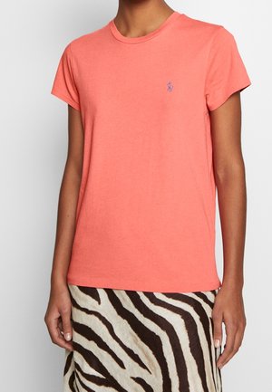 Woman wearing coral short-sleeve t-shirt with small embroidered logo and zebra-striped skirt, standing against plain background.