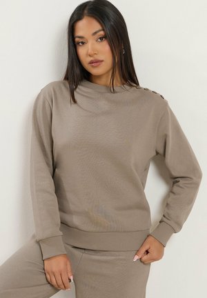 Kvinde med langt mørkt hår iført en taupe sweatshirt og matchende bukser, stående mod en hvid væg med afslappet kropsholdning.