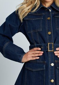 Chaqueta de denim en azul oscuro con dos bolsillos en el pecho, cintura con cinturón y detalles en dorado. Presenta costuras visibles y un diseño estructurado.