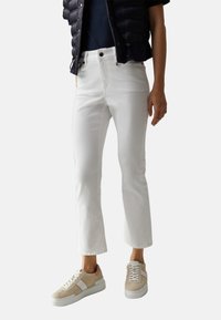 Jean blanc court à la coupe ajustée, associé à des baskets basses beige et blanches. La tenue comprend une veste matelassée foncée et un haut bleu marine.