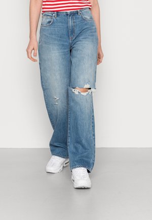 Jeans de perna larga em azul claro com rasgos desfiados, design de cinco bolsos e um acabamento ligeiramente desbotado. Usados com ténis brancos.