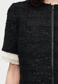 Μαύρο υφασμάτινο κοντομάνικο μπουφάν με φερμουάρ στο μπροστινό μέρος και λευκές στρωματοποιημένες βολάν στα μανίκια.