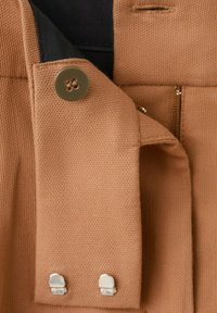 Přiblížení hnědého látkového materiálu ukazujícího rukáv s knoflíky a kovovými háčky a částečný šev s krátkým zipem na oděvu.