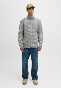 Chemise à carreaux dans des tons bleu clair, beige et vert, associée à un jean bleu et des chaussures beiges. L'individu porte une casquette marron.