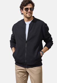 Black zip-up bomber jacket with textured fabric, ribbed cuffs, and hem. Worn over a white T-shirt, paired with light beige pants.