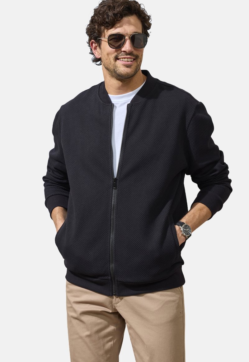 Black zip-up bomber jacket with textured fabric, ribbed cuffs, and hem. Worn over a white T-shirt, paired with light beige pants.
