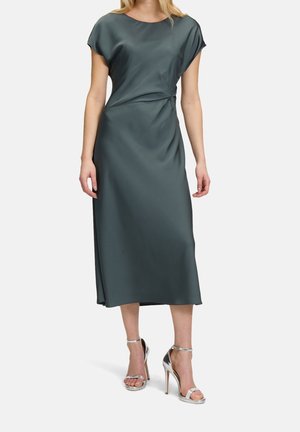 Femme portant une robe midi en satin vert foncé à manches courtes, taille froncée et sandales argentées à talons hauts, debout.
