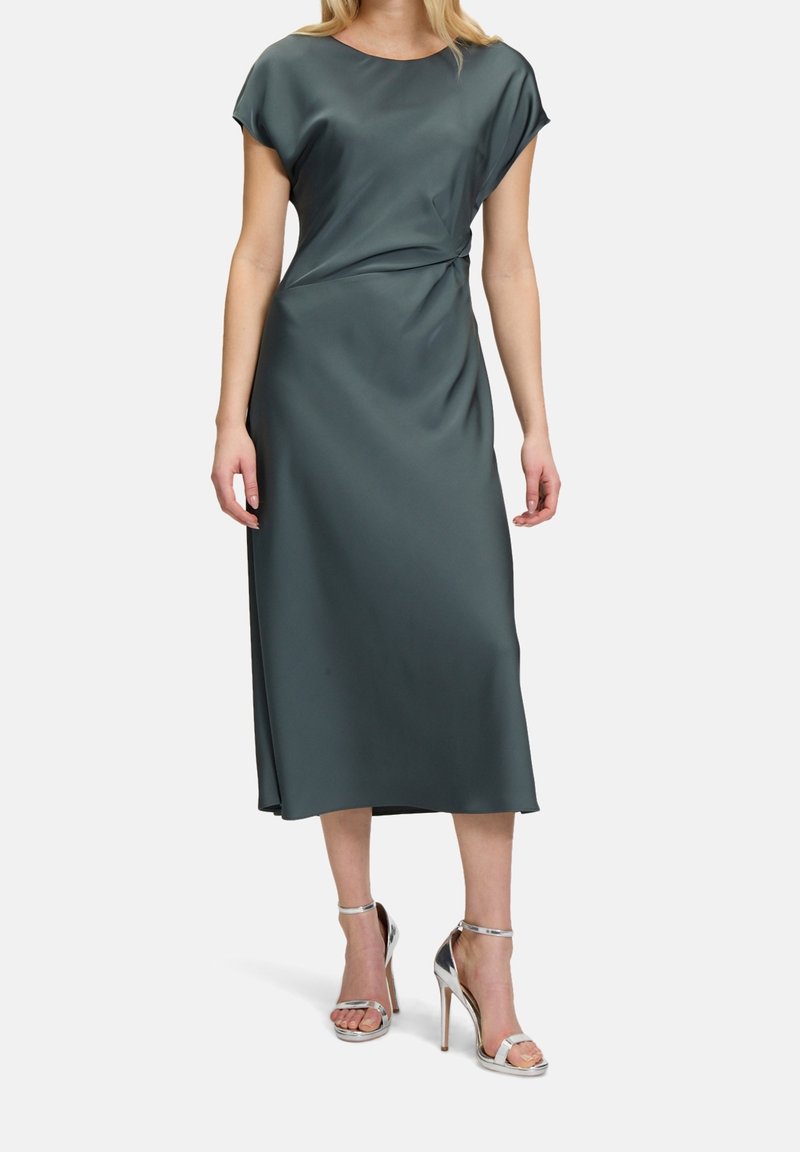 Femme portant une robe midi en satin vert foncé à manches courtes, taille froncée et sandales argentées à talons hauts, debout.