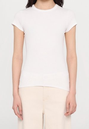 Femme portant un t-shirt blanc uni à manches courtes ajusté et un pantalon beige clair, debout devant un fond neutre.