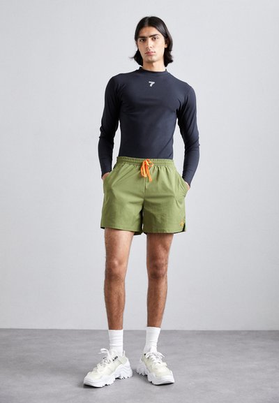 Chemise noire à manches longues, shorts verts avec cordon orange, baskets blanches et chaussettes de cheville ; style athlétique décontracté.