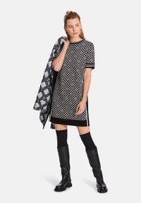 Schwarz-weiß gemustertes Kleid mit kurzen Ärmeln, begleitet von einem gemusterten Schal. Das Model trägt kniehohe schwarze Stiefel und schwarze Kniestrümpfe.