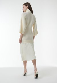 Crèmekleurige, ribbelgebreide jurk met een hoge kraag en wijde mouwen, gelaagd over een geplooide, vloeiende rok. Draagt witte hoge hakken.