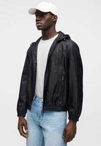 Μαύρο αντιανεμικό μπουφάν με υφή και σχέδιο, που διαθέτει κουκούλα, τσέπες με φερμουάρ και ελαστικό τελείωμα. Φοράται πάνω από γκρι πουκάμισο και ανοιχτόχρωμο τζιν.