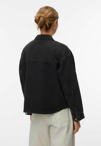 Crna traper jakna opuštenog kroja, s velikim ovratnikom i manžetama na kopčanje. Ima šav na stražnjem dijelu i sirovi rub. Nošena preko svijetlih hlača.