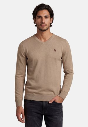 Mann mit dunklen Haaren, der einen beigen V-Ausschnittpullover mit kleinem gesticktem Logo auf der Brust und schwarze Jeans trägt, eine Hand in der Tasche.