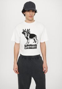 Weißes Baumwoll-T-Shirt mit einem schwarzen Grafikdruck von zwei Deutschen Schäferhunden und dem Text "CERBERUS" darunter, kombiniert mit dunklen Hosen und einer Mütze.