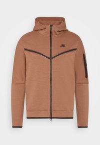 Hnědá zipová mikina s kapucí, černé akcenty na ramenou a vpředu, strukturovaná tkanina a zipová kapsa na levém rukávu.