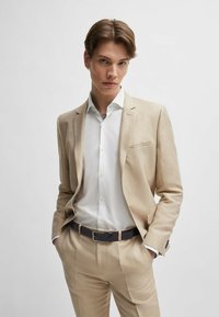 Lichtbeige kostuum gemaakt van gestructureerde stof, met een notch-revers en een enkele zak, gecombineerd met een witte blouse en een donkere riem.