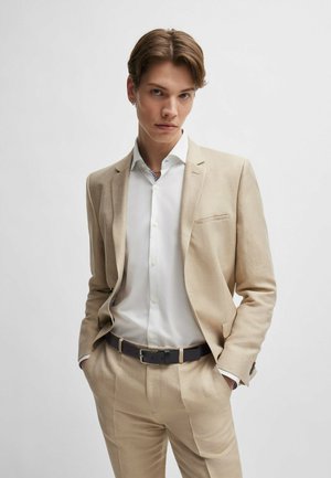 Traje beige claro hecho de tela texturizada, que presenta una solapa muescada y un bolsillo simple, combinado con una camisa blanca y un cinturón oscuro.