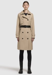 Beige double-breasted trench coat with a black belt, large collar, and button accents. Worn over black leggings and ankle boots.
