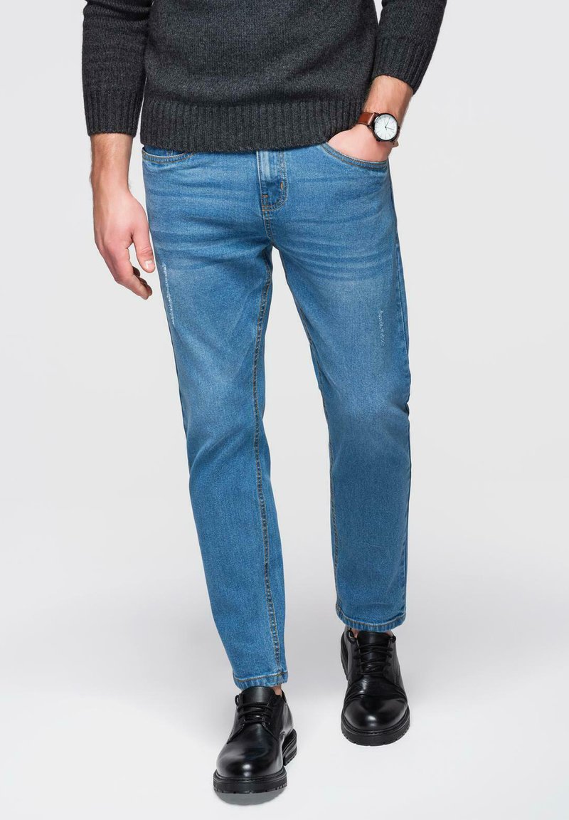 Jean en denim bleu clair avec une coupe droite, taille mi-haute et détails délavés. Associé à des chaussures en cuir noires.