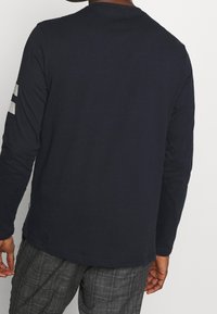 Man i en långärmad mörkblå skjorta med reflexremsor på vänster ärm och grå rutiga byxor, visad från baksidan.