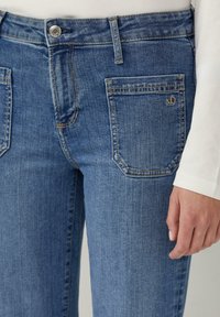 Blaue Jeans aus Denim mit glatter Textur und subtilen Verblassungen, ausgestattet mit zwei Gesäßtaschen und einem kleinen Logo auf der rechten Tasche.