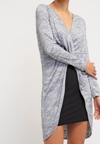 Šedý melírovaný top s dlouhým rukávem, hlubokým výstřihem do V a zkrouceným předním designem, v kombinaci s černou přiléhavou sukní, která ukazuje vrstvený vzhled.