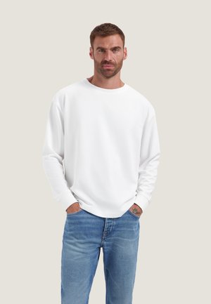 Uomo con la barba che indossa una maglietta bianca semplice a maniche lunghe e jeans blu, in piedi con le mani nelle tasche davanti a uno sfondo chiaro.