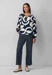 Haut à manches longues bleu marine et crème avec des motifs circulaires et un col à nouer, associé à un pantalon large gris et des mocassins crème.