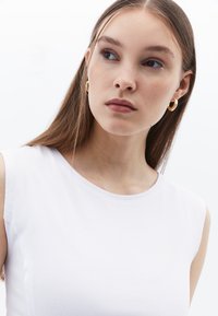 Bílý žebrovaný top bez rukávů s kulatým výstřihem. Model má na sobě zlaté kruhové náušnice a hladké vlasy částečně stažené. Čisté, minimalistické pozadí.