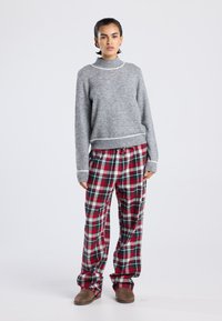 Jongere staat gekleed in een grijze trui, een rood-groene ruitbroek en een paar bruine pantoffels tegen een effen witte achtergrond.