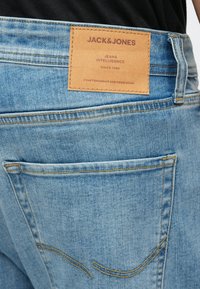 Lichtblauwe spijkerbroek van denim met een leren patch met merknaam en informatie. Verweerde textuur met gele stikaccenten.