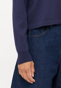 Pullover in maglina blu navy con polsini e orlo a coste, abbinato a jeans di denim scuri. Dettagli visibili sulla manica e sulla mano.